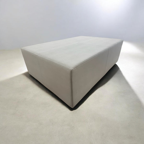 A modern, low-profile rectangular ottoman or coffee table upholstered in a light beige fabric, shown in a minimalist studio setting with a concrete floor and white walls.