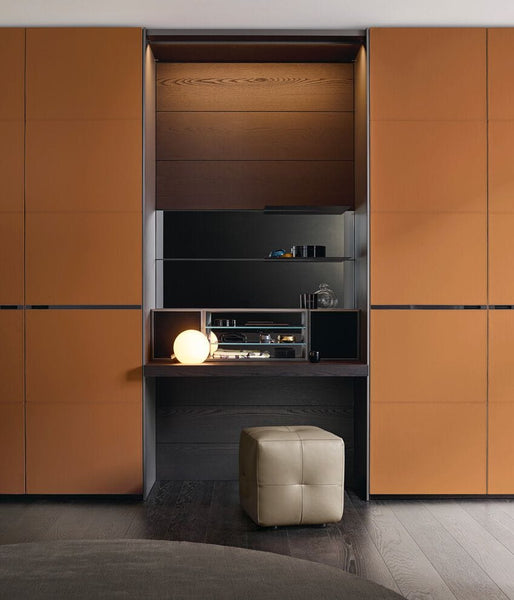 A modern built-in cabinetry and desk unit with warm orange-brown panel doors, dark wood interior shelving, a lit spherical lamp, and a small tufted cream ottoman in a styled interior setting.