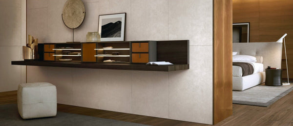 A modern wall-mounted floating desk in dark wood and warm brown tones, styled in a minimalist bedroom interior with a white bed, wood paneling, and a white ottoman.