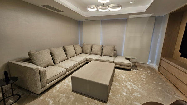 A modern U-shaped sectional sofa in a warm beige fabric, arranged in a home theater or living room setting with a matching rectangular ottoman, textured carpet, and ambient wall lighting.