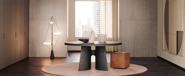 A modern round dining table with a dark stone pedestal base, styled in a minimalist dining room with a light brown leather pouf, a geometric floor lamp, and a round beige rug.