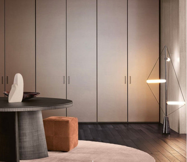 A modern interior scene featuring a large wall of tall, minimalist wardrobe doors in a warm taupe-beige finish, set against dark wood flooring and a light-colored rug. In the foreground, a dark round table with a textured gray base, a small brown cube ottoman, and a white sculptural vase are visible, alongside a contemporary geometric floor lamp on the right.